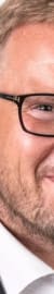 Porträt eines Mannes mit Brille, Anzug und weißem Hemd vor weißem Hintergrund.