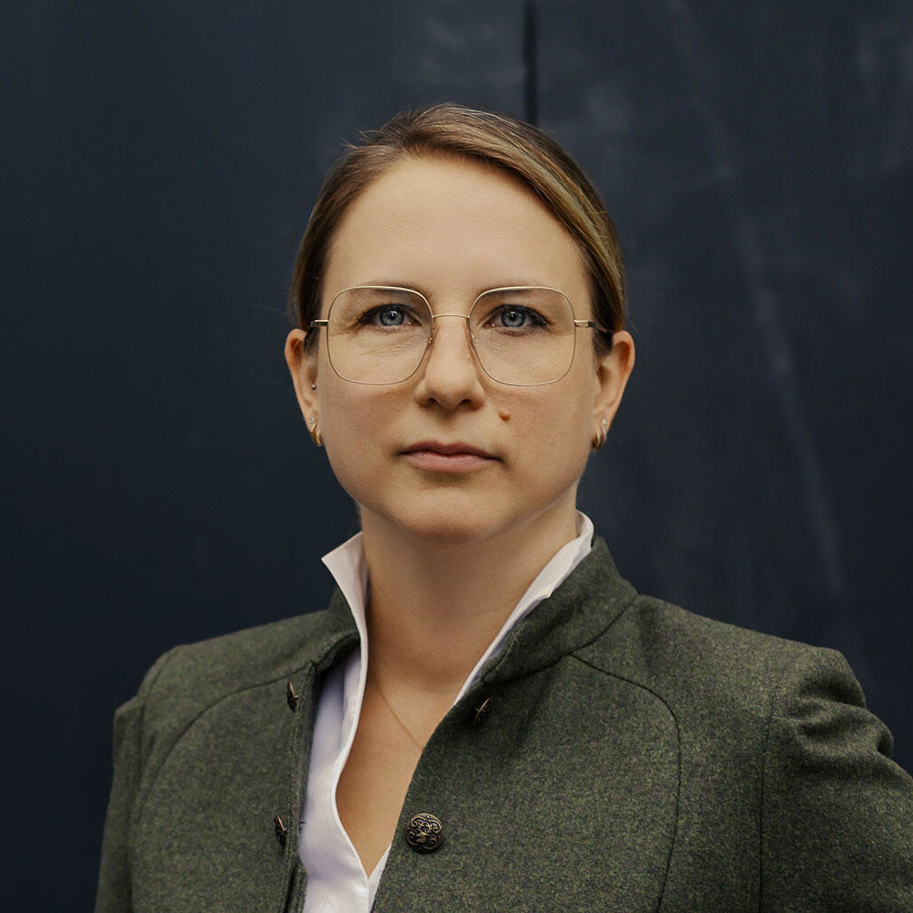 Frau mit Brille und grauer Jacke vor dunklem Hintergrund.