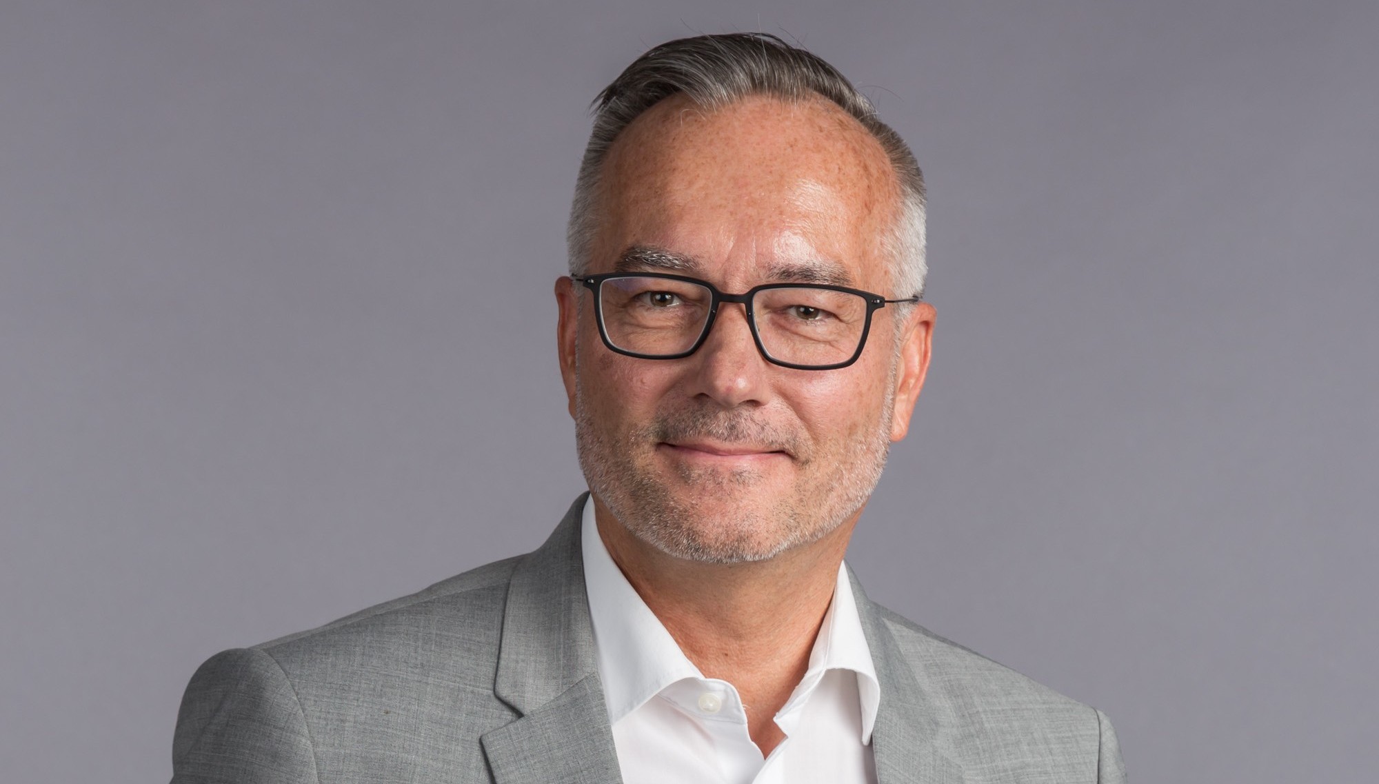 Porträt eines Mannes in einem grauen Anzug und weißen Hemd, mit Brille, vor einem grauen Hintergrund.