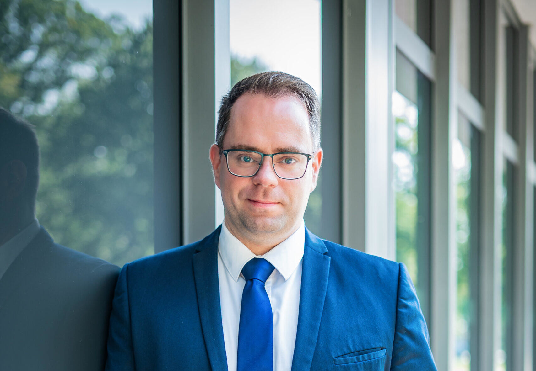 Geschäftsmann in blauer Anzugjacke und Krawatte, stehend neben Glasfenster, draußen grüne Bäume sichtbar.