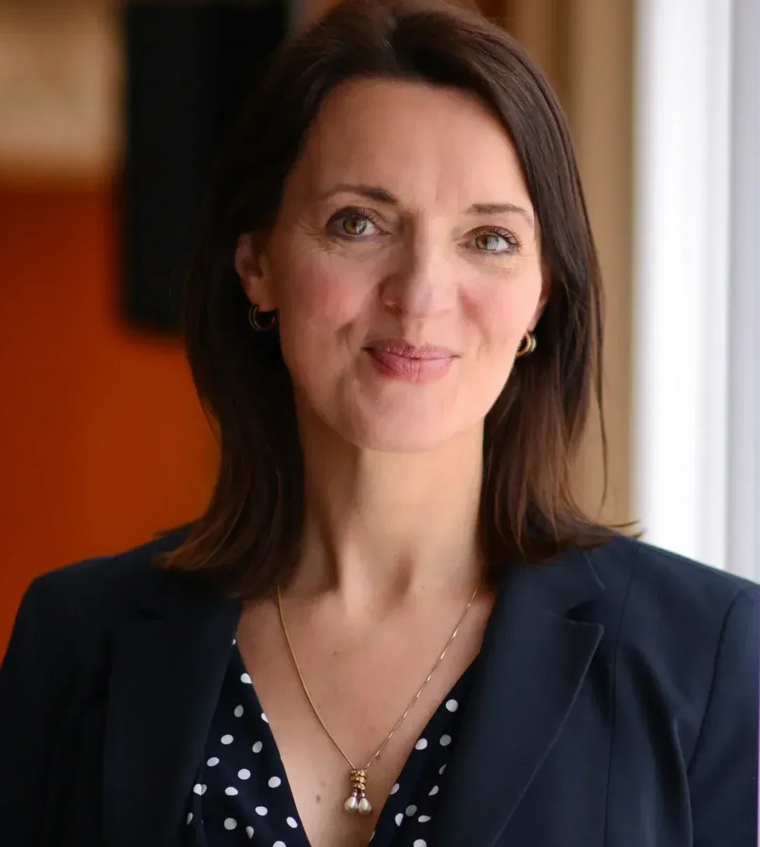 Frau in dunkler Blazer und Polka-Dot-Bluse vor einem Fenster mit weißen Vorhängen.