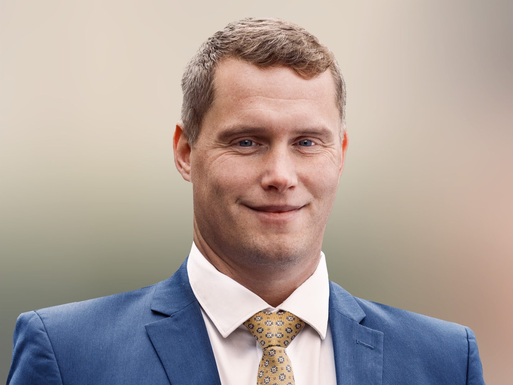 Mann in blauer Anzug mit weißem Hemd und gelber Krawatte, lächelnd vor neutralem Hintergrund.