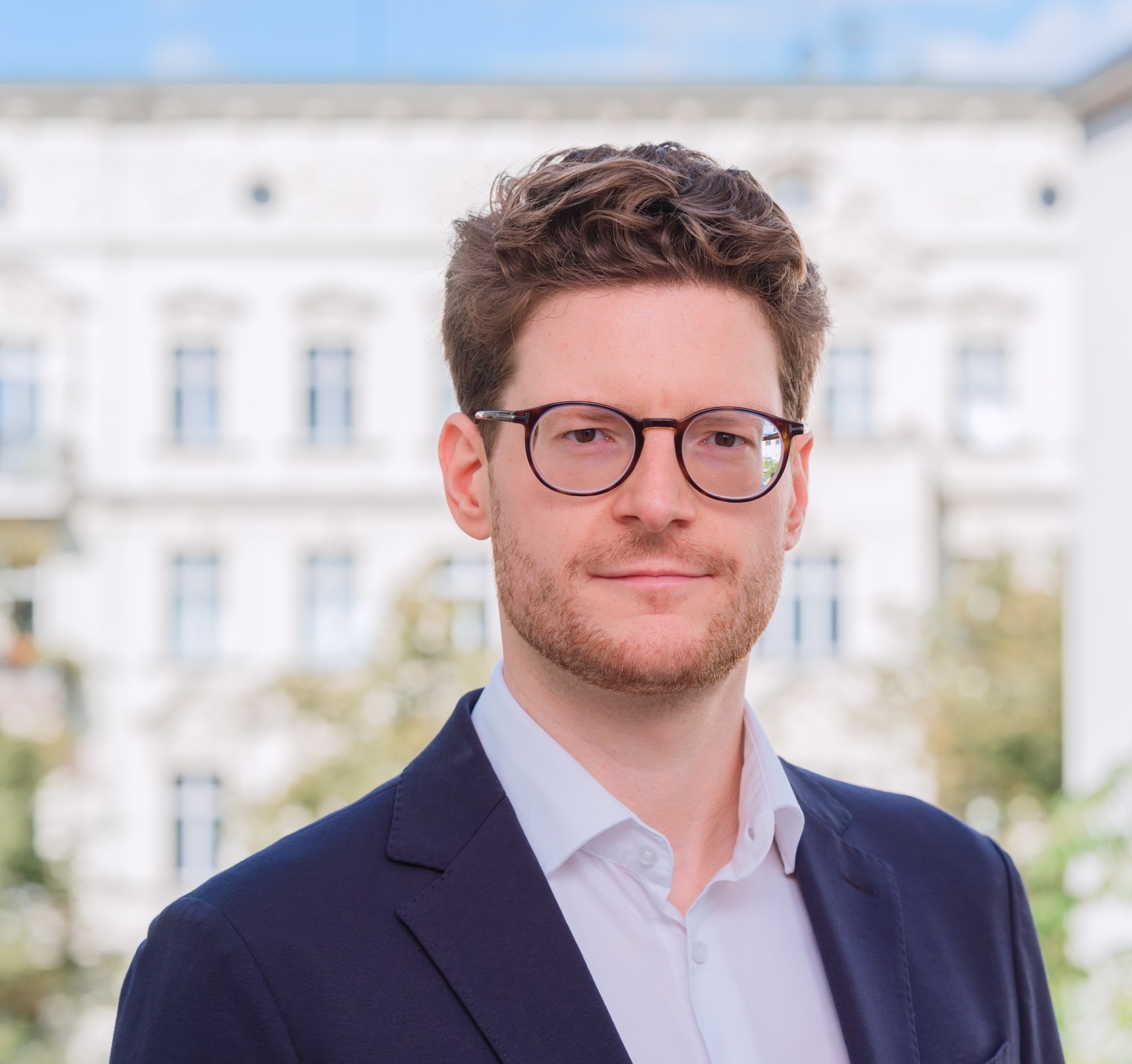 Junger Mann in formeller Kleidung mit Brille vor einem weißen Gebäude.