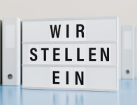 Weiße Aktenordner und ein Schild mit der Aufschrift "WIR STELLEN EIN" auf einem blauen Tisch.