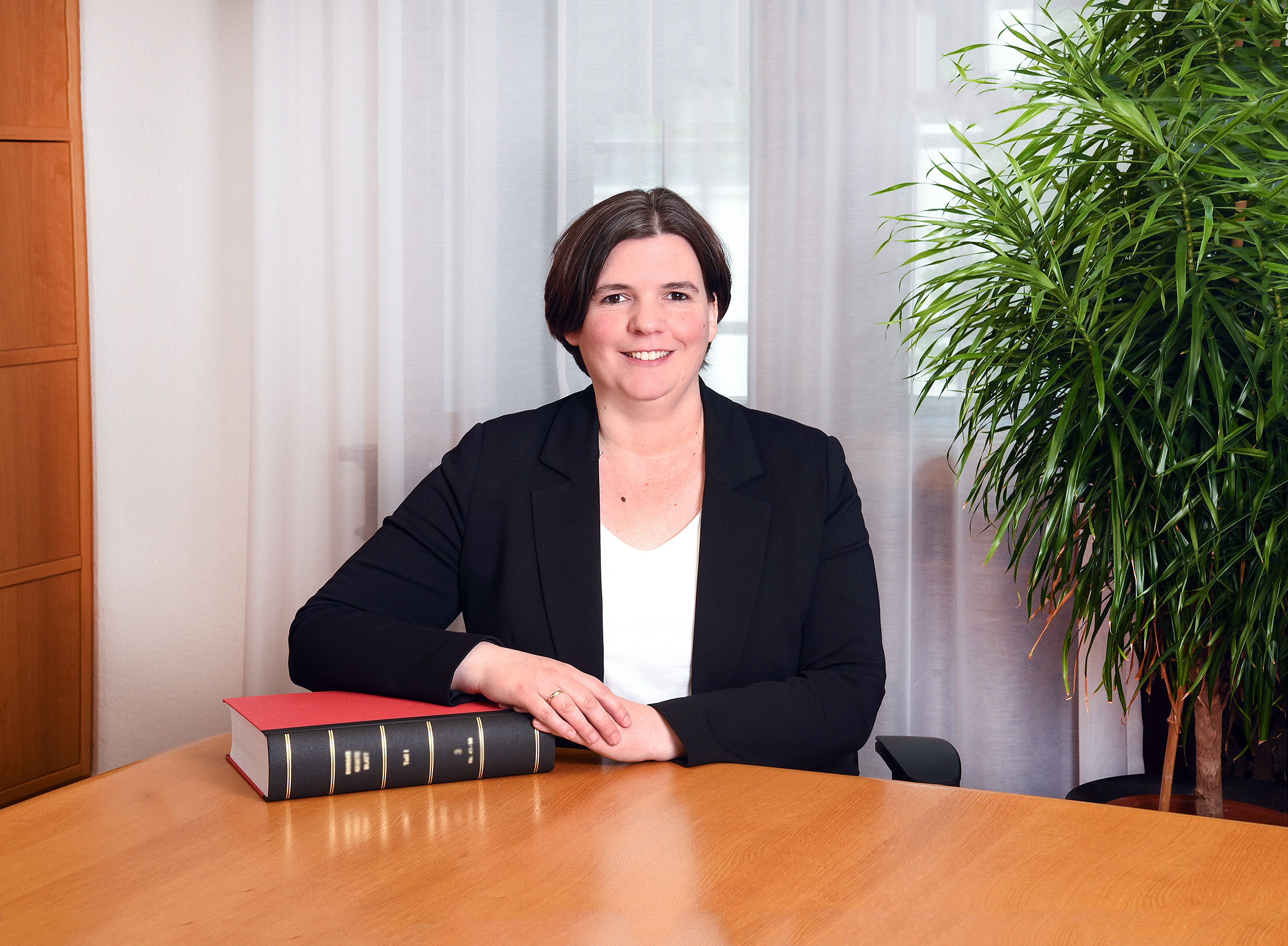 Frau sitzt an einem Tisch im Büro, trägt eine schwarze Jacke und hat kurze dunkle Haare.