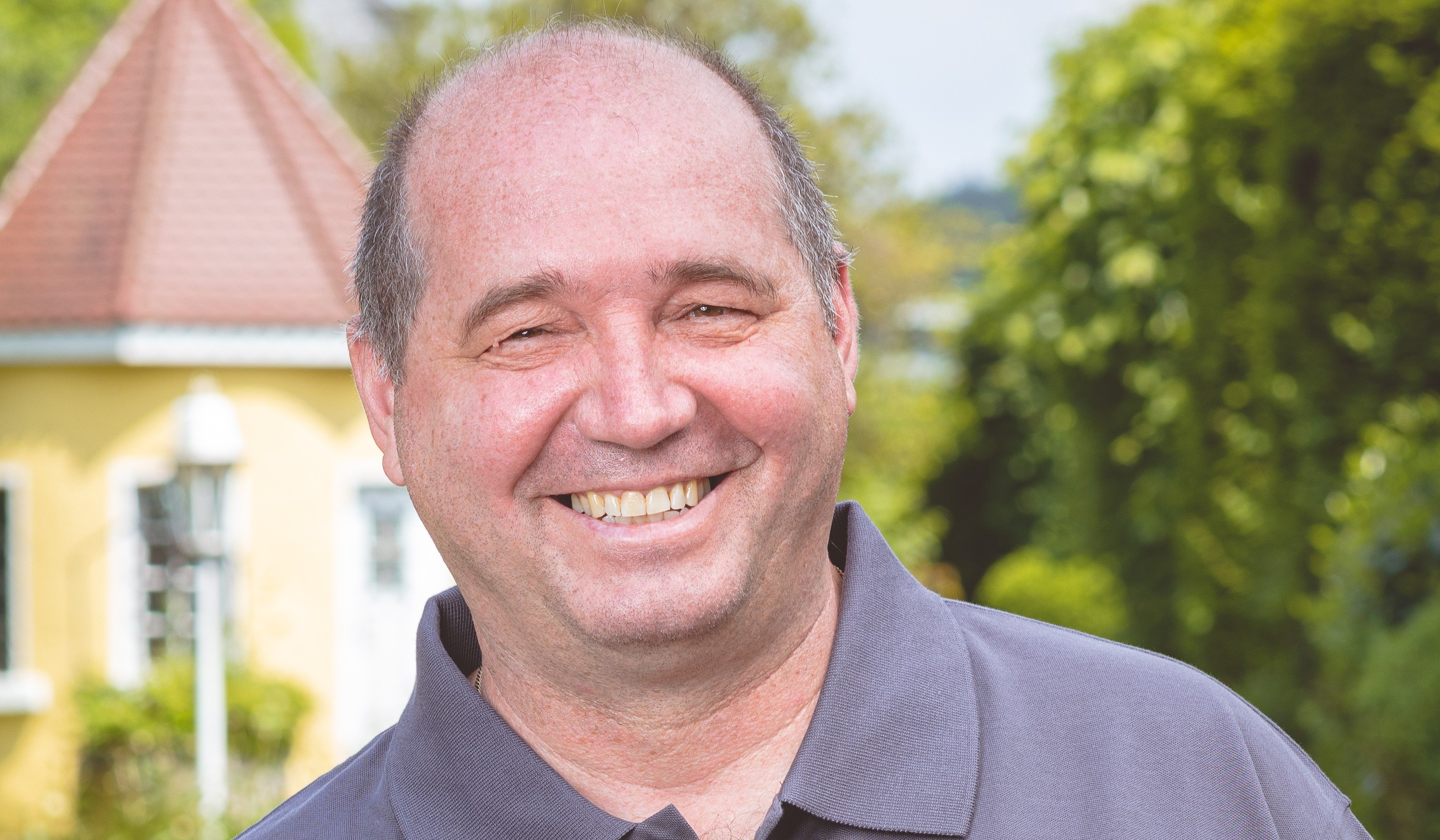 Lächelnder Mann in einem grauen Polo-Shirt mit "Immobilienwelt KRESS" Logo, im Freien vor einem gelben Haus und grünen Bäumen.