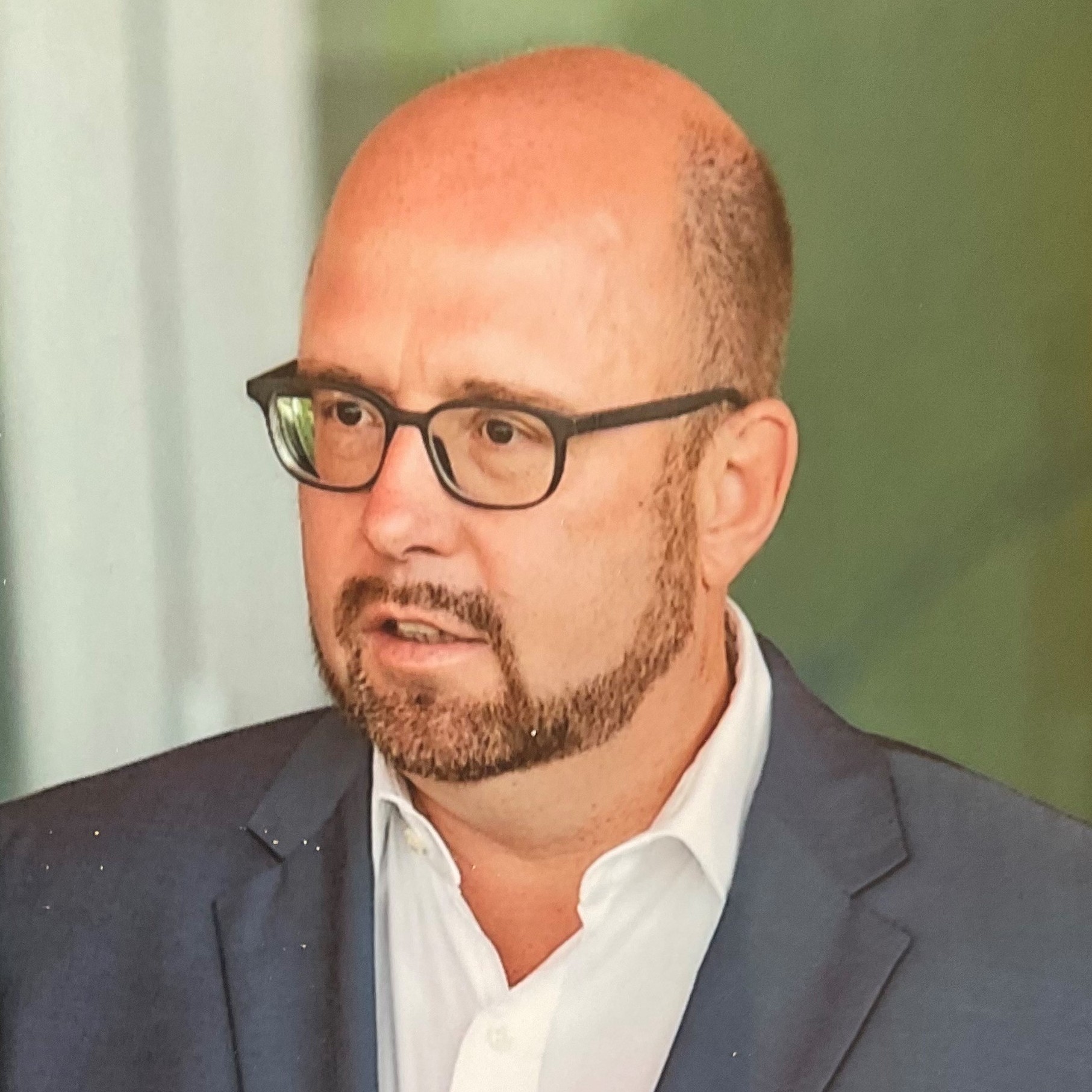 Ein Mann mit Brille, Bart und Glatze trägt einen dunklen Anzug und eine weiße Hemd, im Hintergrund ist eine grüne Wand.