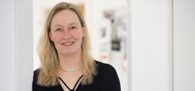 Lächelnde Frau mit blonden Haaren in einem schwarzen Oberteil vor einem weißen Hintergrund.