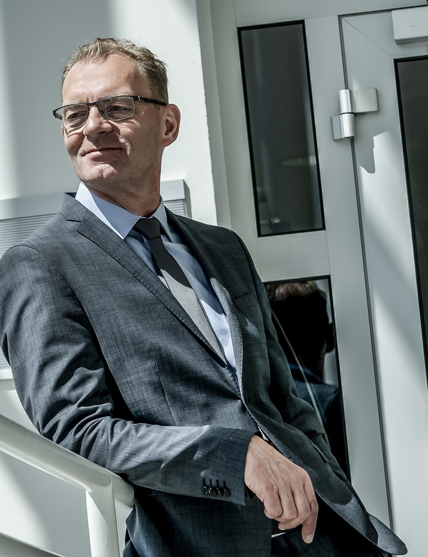 Ein Mann in einem grauen Anzug mit Krawatte steht draußen vor einem Gebäude, lehnt an einem Geländer und schaut nach rechts.