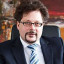 Ein Mann in Anzug mit Brille sitzt an einem Schreibtisch vor einem bunten Gemälde.