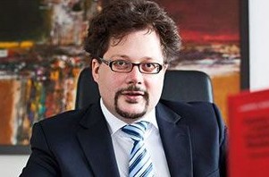 Ein Mann in Anzug mit Brille sitzt an einem Schreibtisch vor einem bunten Gemälde.