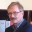 Ein Mann mit Brille und grauem Haar steht vor einem Bücherregal mit roten und weißen Büchern.