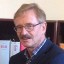 Ein Mann mit Brille und grauem Haar steht vor einem Bücherregal mit roten und weißen Büchern.