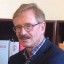 Ein Mann mit Brille und grauem Haar steht vor einem Bücherregal mit roten und weißen Büchern.