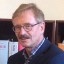 Ein Mann mit Brille und grauem Haar steht vor einem Bücherregal mit roten und weißen Büchern.