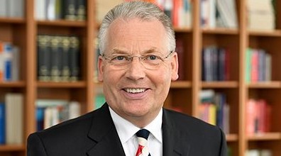 Ein älterer Mann in formeller Kleidung vor einem Bücherregal.