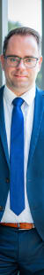 Geschäftsmann in blauer Anzugjacke und Krawatte, stehend neben Glasfenster, draußen grüne Bäume sichtbar.
