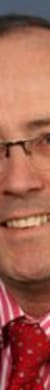 Porträt eines Mannes mit Brille, dunklem Anzug, roter Krawatte und gestreiftem Hemd vor blauem Hintergrund.