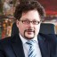 Ein Mann in Anzug mit Brille sitzt an einem Schreibtisch, umgeben von roten Aktenordnern.
