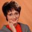 Eine Frau mit kurzen dunklen Haaren, Brille und einem grauen Blazer, die lächelnd in die Kamera schaut, vor einem orangefarbenen Hintergrund.