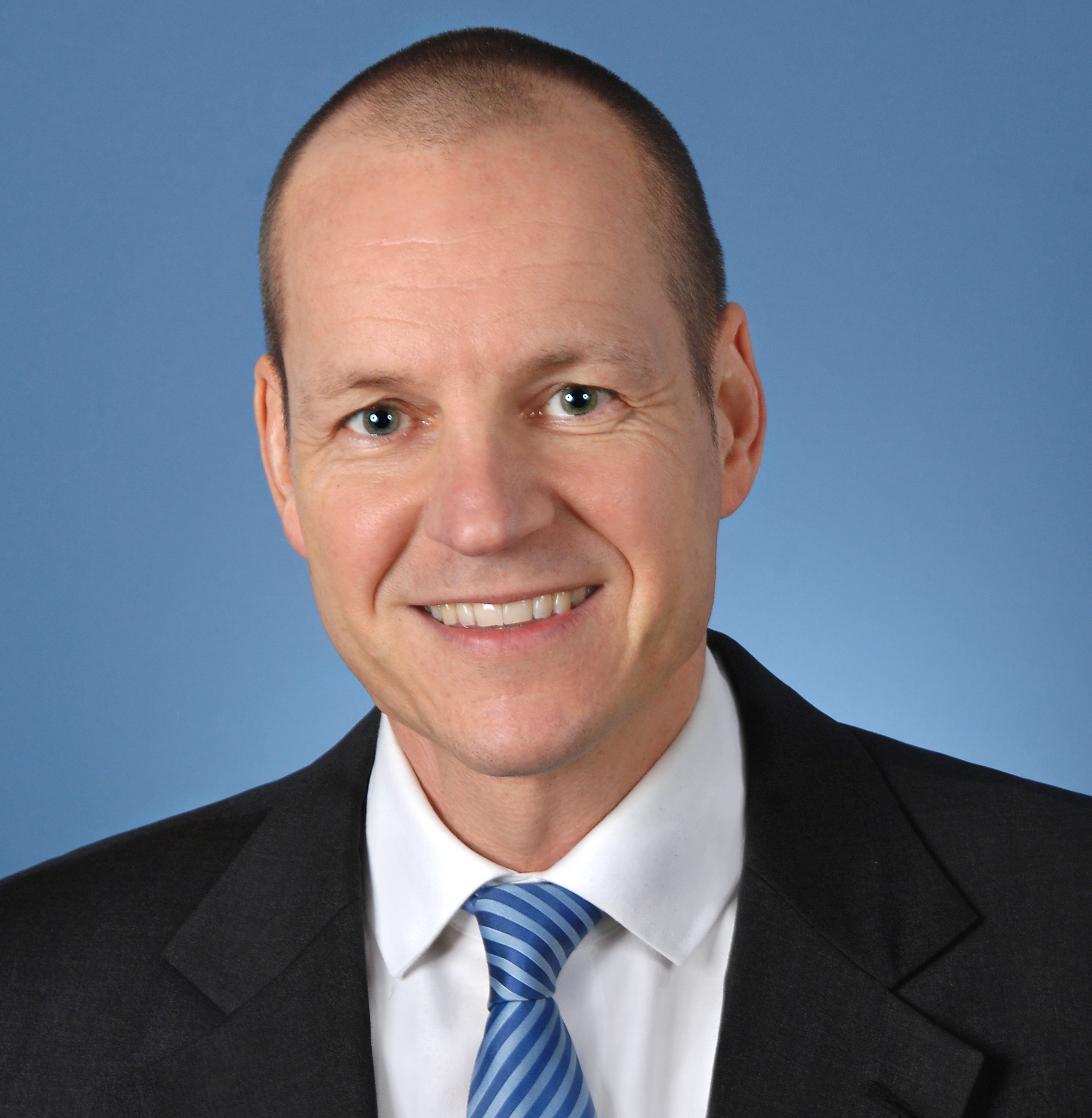 Porträt eines lachenden Mannes in formeller Kleidung vor einem blauen Hintergrund.