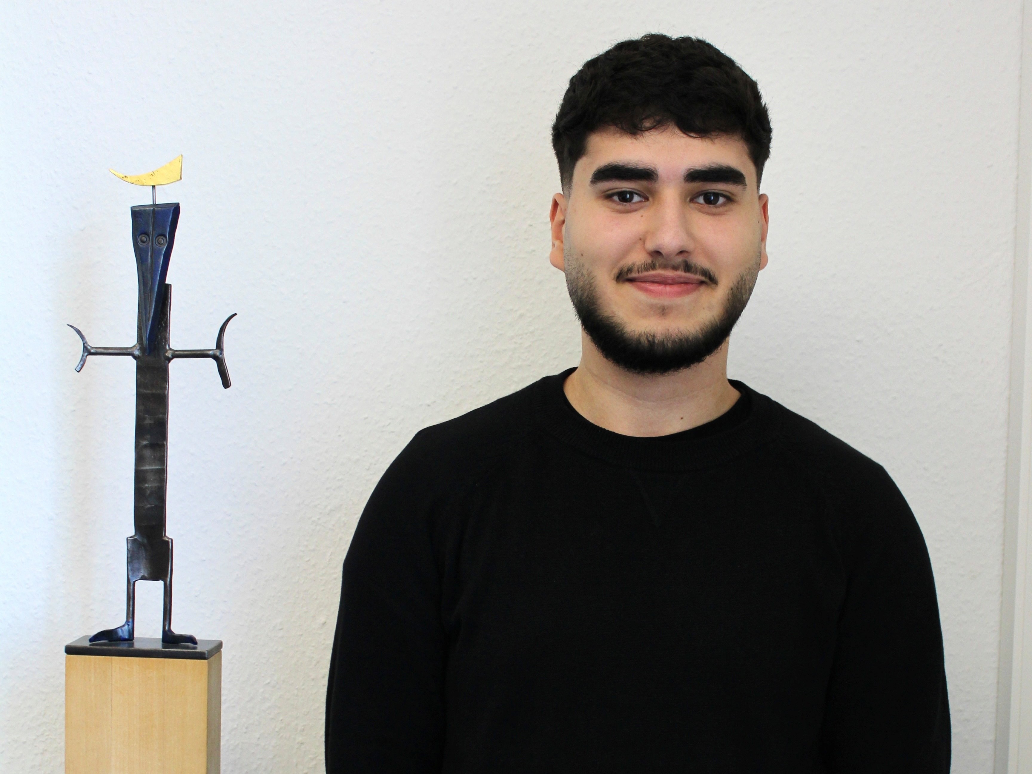 Junge Person mit dunklem Haar und Bart neben einer Skulptur auf einem Holzsockel.