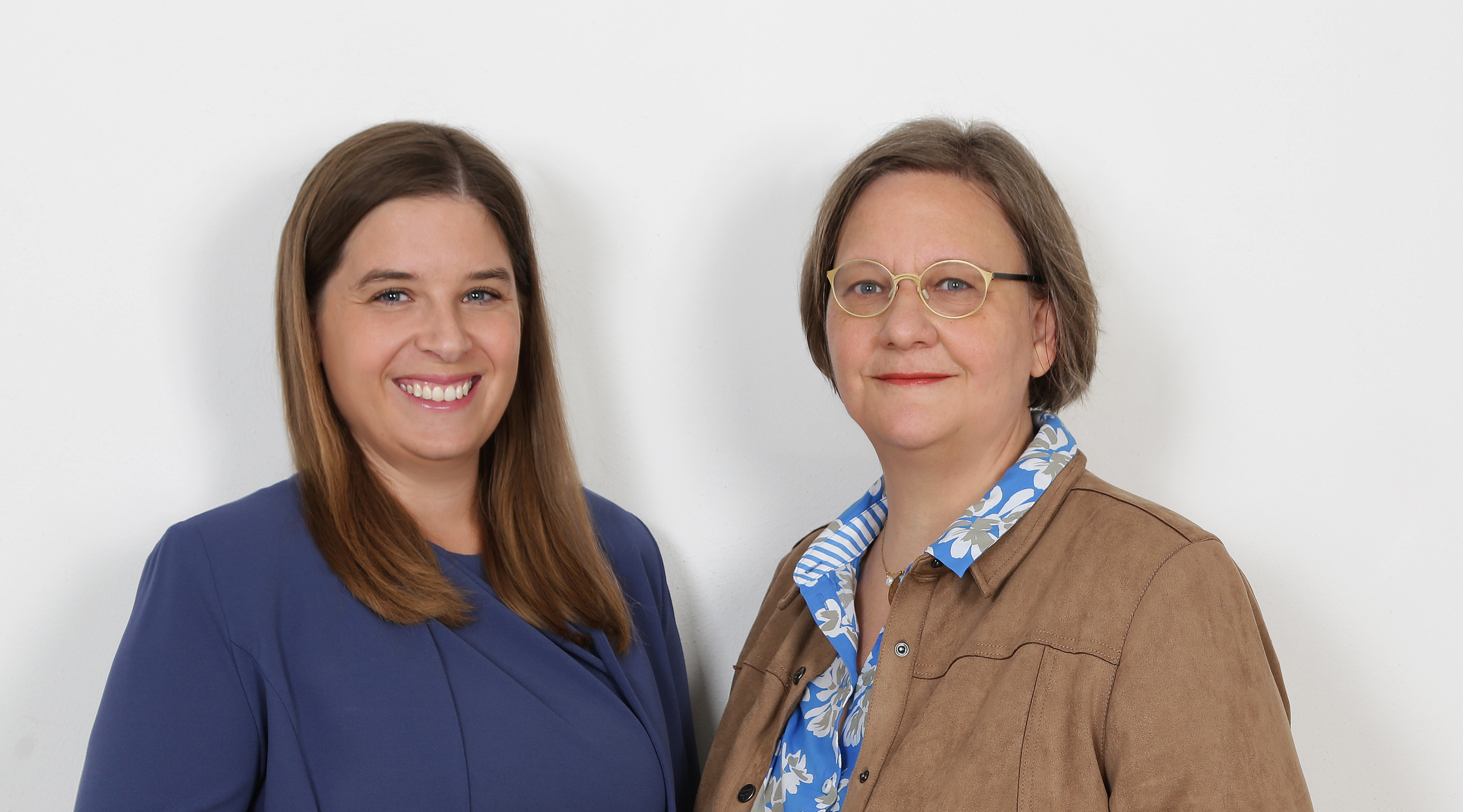 Zwei Frauen stehen nebeneinander vor weißem Hintergrund, lächeln und schauen in die Kamera.