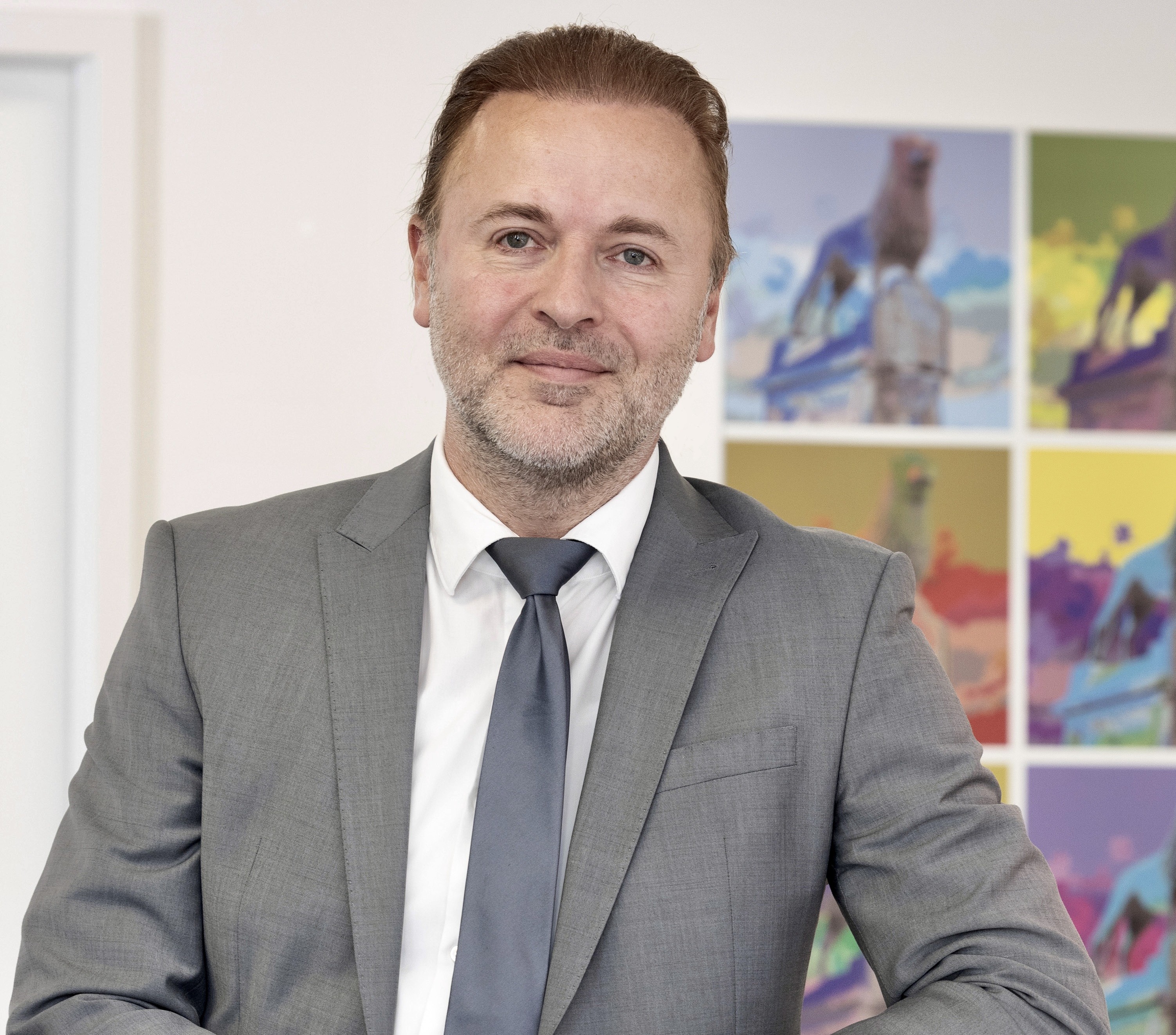 Ein Mann in einem grauen Anzug steht vor einem weißen Schrank und lächelt. Im Hintergrund sind bunte Pop-Art-Bprints eines Hundes an der Wand zu sehen.