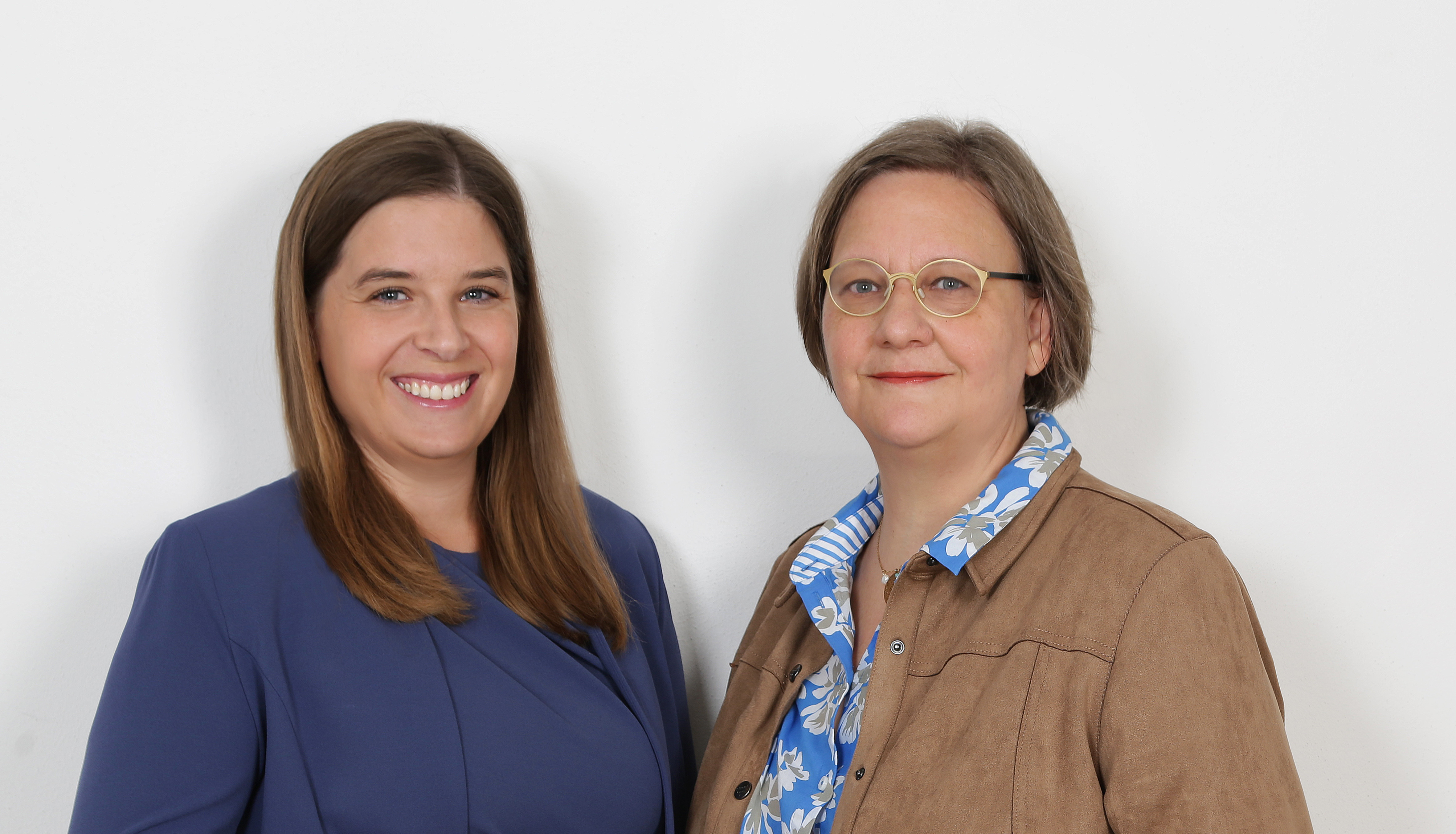 Zwei Frauen stehen nebeneinander vor weißem Hintergrund, lächeln und schauen in die Kamera.