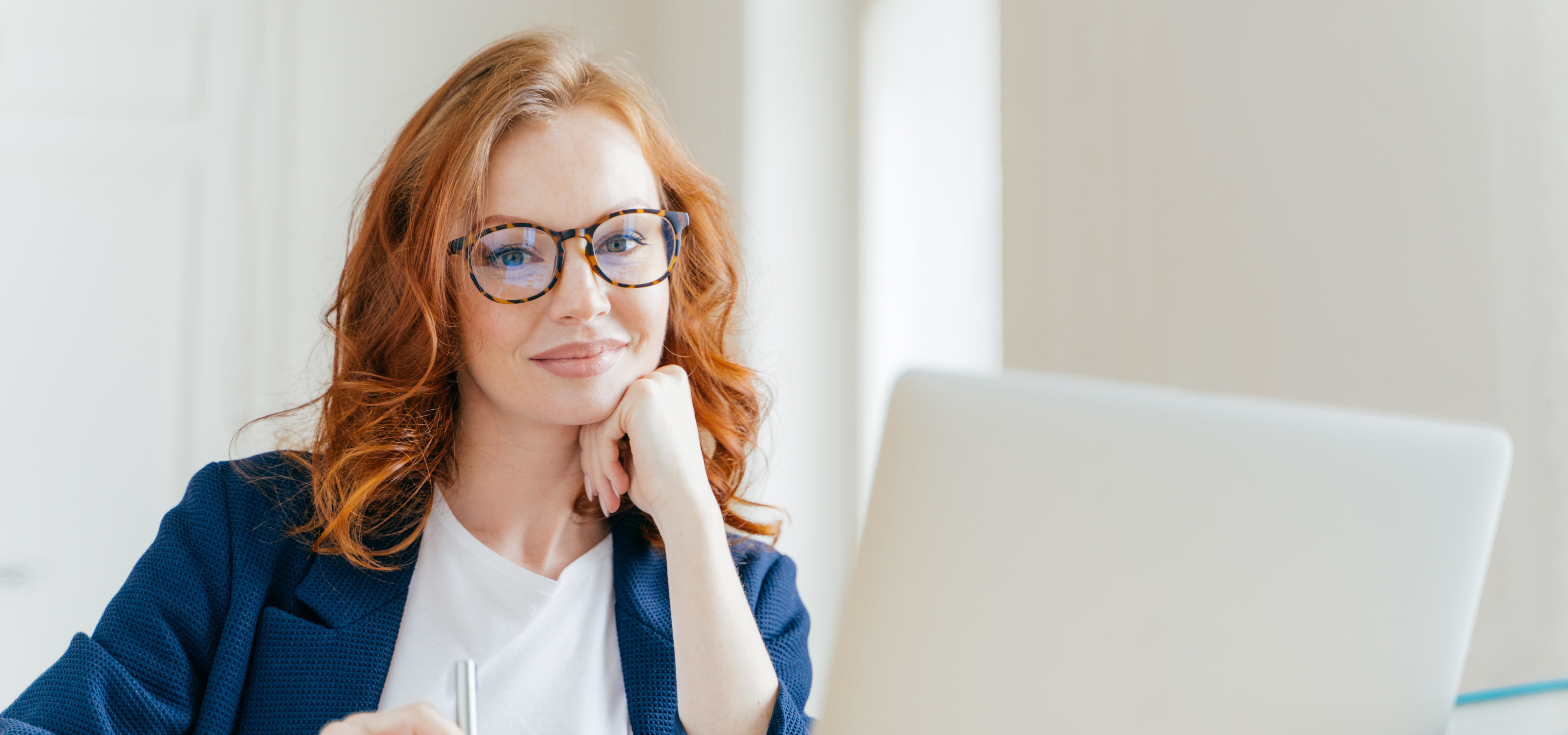 Frau mit roten Haaren und Brille arbeitet an einem Laptop im Büro.