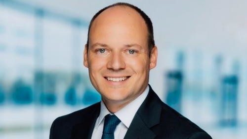 Ein Mann in einem dunklen Anzug und Krawatte lächelt vor einem modernen Bürogebäude.