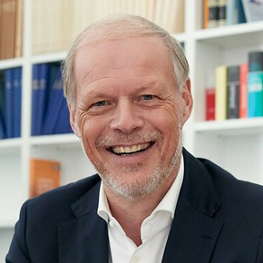 Lächelnder Mann in Anzug mit Smartphone im Büro vor Bücherregal.