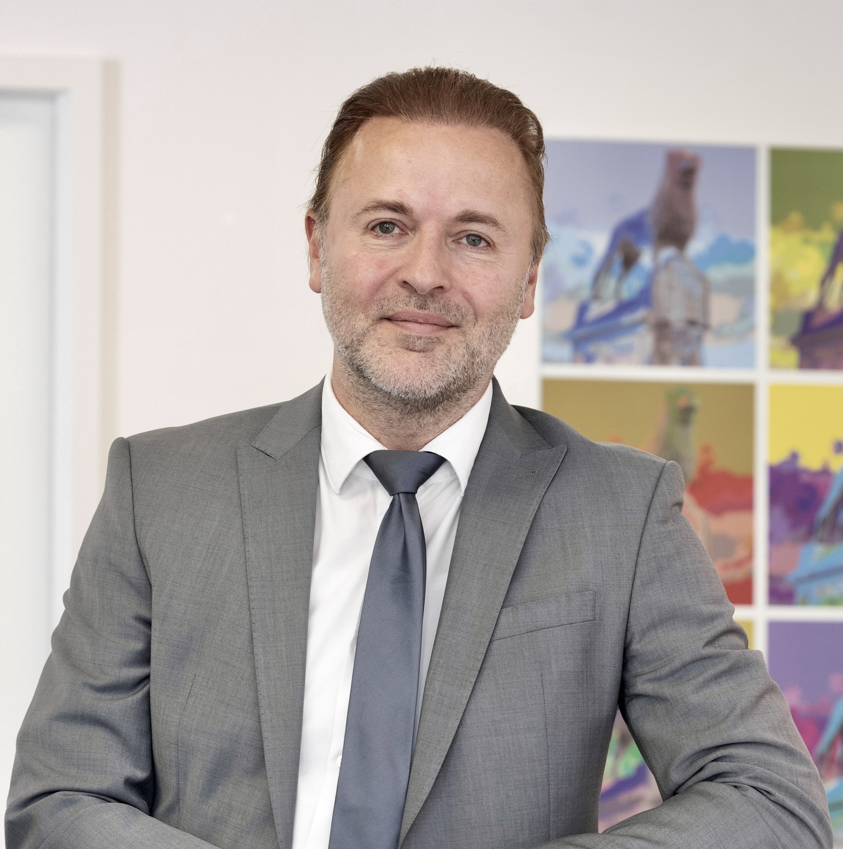 Ein Mann in einem grauen Anzug steht vor einem weißen Schrank und lächelt. Im Hintergrund sind bunte Pop-Art-Bprints eines Hundes an der Wand zu sehen.