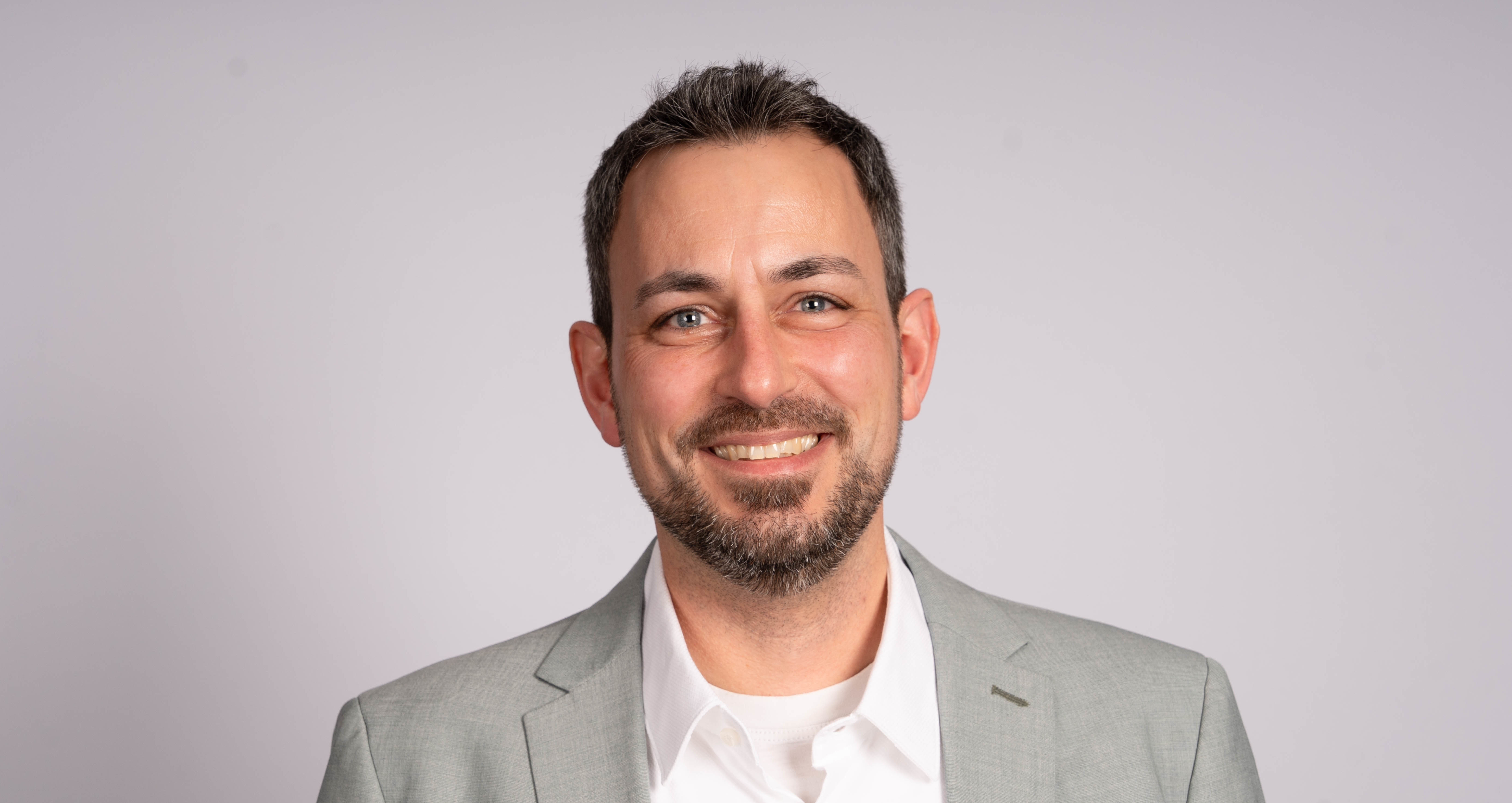 Lächelnder Mann in einem hellgrauen Anzug und weißem Hemd vor neutralem Hintergrund.