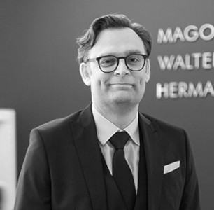 Ein Mann in formeller Kleidung mit Brille vor einer Wand mit der Aufschrift "MAGOLD, WALTER & HERMANN".