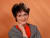 Eine Frau mit kurzen dunklen Haaren, Brille und einem grauen Blazer, die lächelnd in die Kamera schaut, vor einem orangefarbenen Hintergrund.