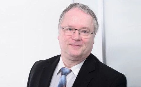 Ein Mann in Anzug und Krawatte steht vor einer weißen Wand.