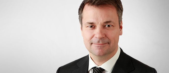 Ein Mann in einem schwarzen Anzug und Krawatte vor hellem Hintergrund.