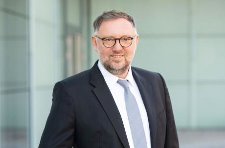 Ein Mann in einem schwarzen Anzug mit Krawatte und Brille steht vor einem modernen Gebäude.