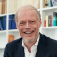 Lächelnder Mann in Anzug mit Smartphone im Büro vor Bücherregal.