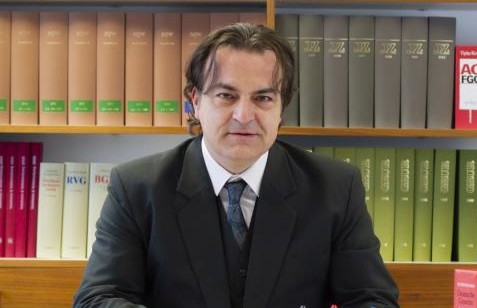 Ein Mann in Anzug sitzt an einem Schreibtisch vor einem Bücherregal mit bunten Aktenordnern.