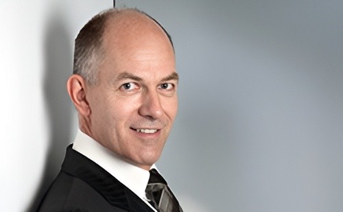 Porträt eines Mannes in formeller Kleidung, lächelnd, vor einem neutralen Hintergrund.