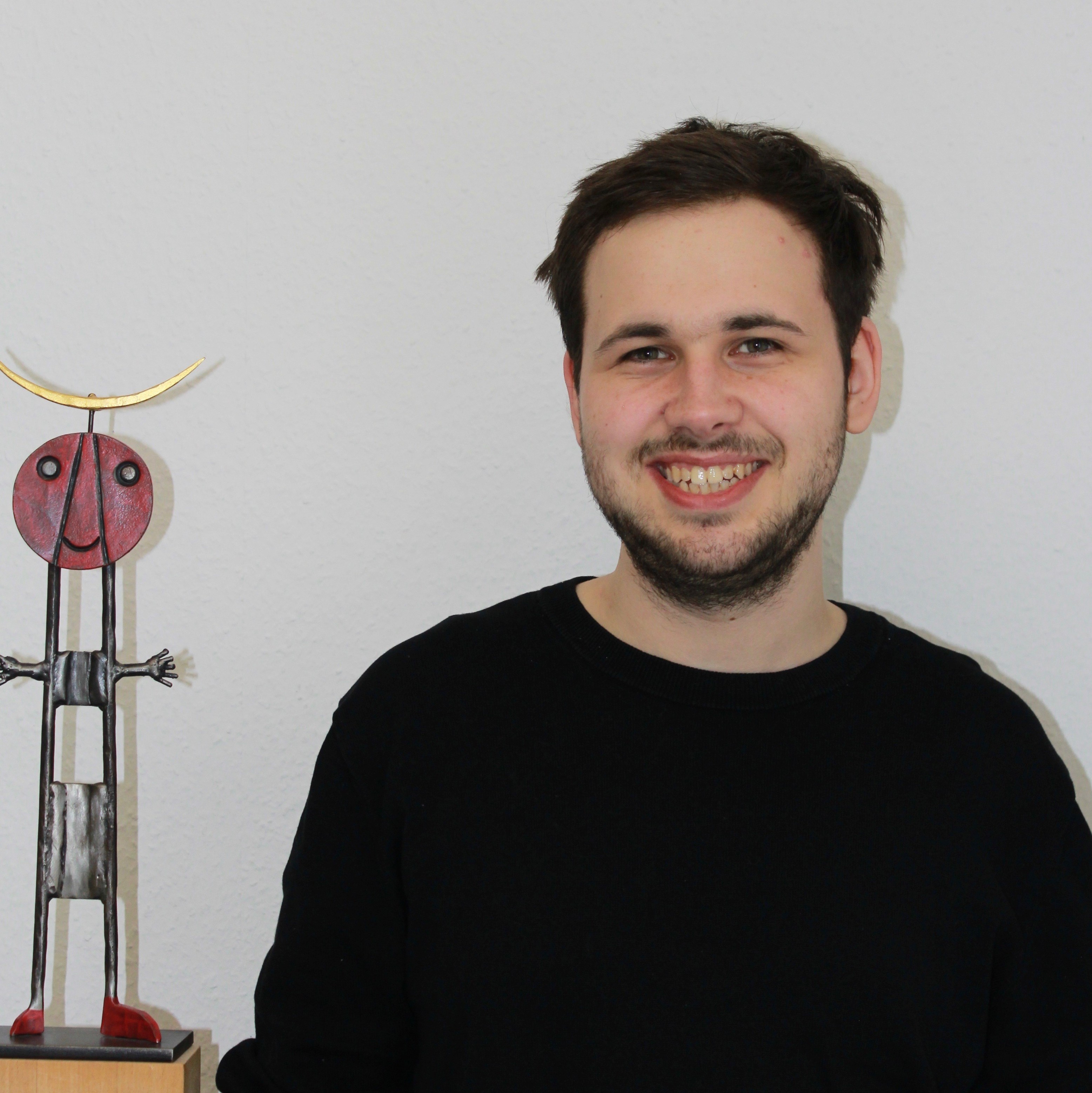 Lächelnder junger Mann neben einer Skulptur auf einem Holzsockel.