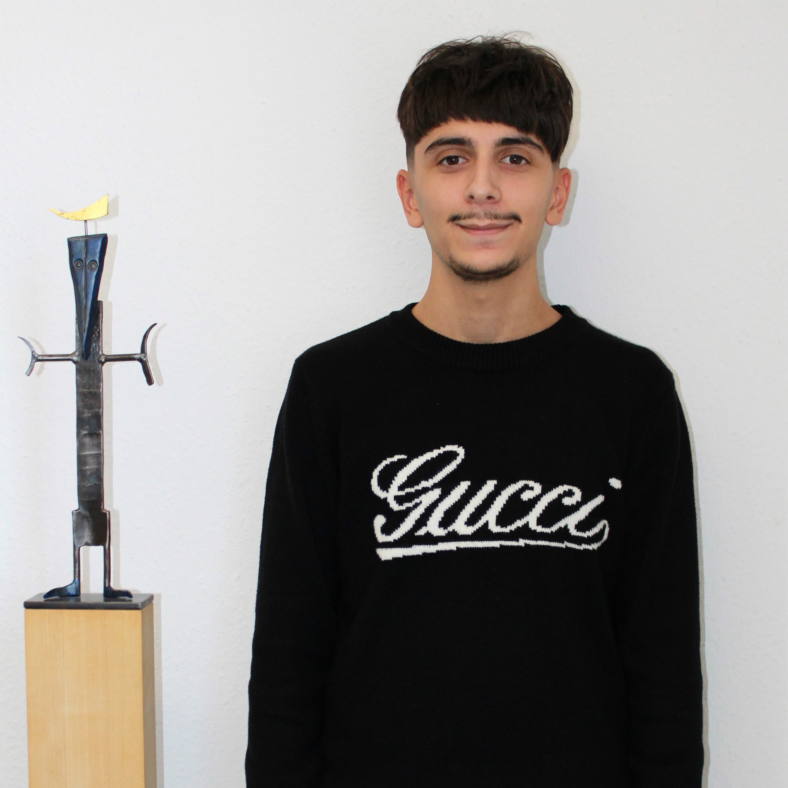 Junger Mann steht neben einer Skulptur eines Schwertes auf einem Holzsockel vor einer weißen Wand.