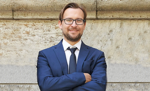 Geschäftsmann in Anzug und Brille vor einer Steinmauer, lächelnd mit verschränkten Armen.