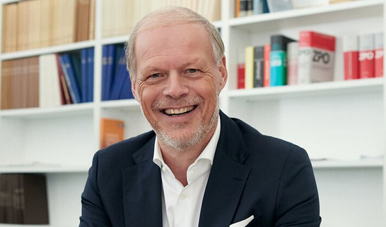 Lächelnder Mann in Anzug mit Smartphone im Büro vor Bücherregal.
