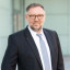 Ein Mann in einem schwarzen Anzug mit Krawatte und Brille steht vor einem modernen Gebäude.