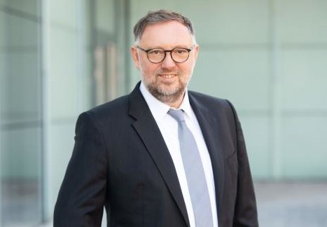 Ein Mann in einem schwarzen Anzug mit Krawatte und Brille steht vor einem modernen Gebäude.