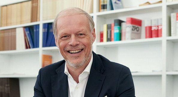 Lächelnder Mann in Anzug mit Smartphone im Büro vor Bücherregal.