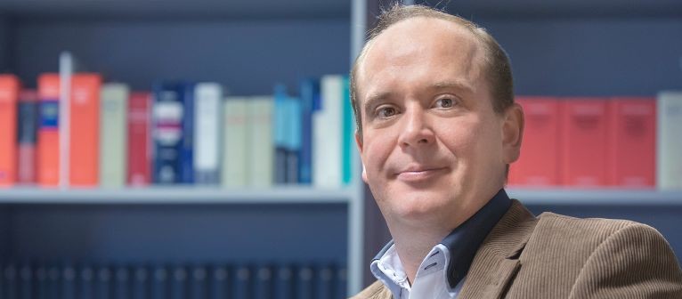 Geschäftsmann sitzt an einem Tisch in einem Büro mit Bücherregalen im Hintergrund.